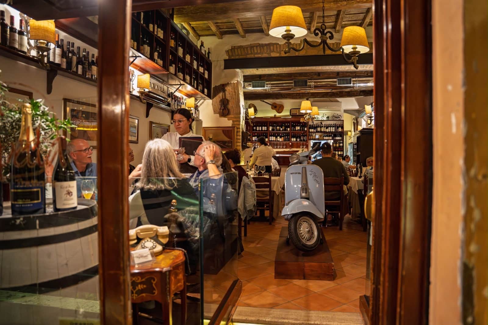 Sala interna vista dalla porta a vetri, con scaffali di bottiglie di vino e uno scooter d'epoca celeste esposto tra i tavoli.