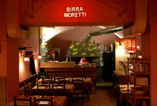 Sala con forno a legna in muratura, insegna al neon rossa con scritta Birra Moretti e tavoli apparecchiati in primo piano.