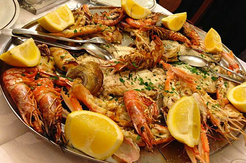 Vassoio metallico con gamberoni, calamari e pesce grigliato, guarnito con spicchi di limone e prezzemolo tritato.
