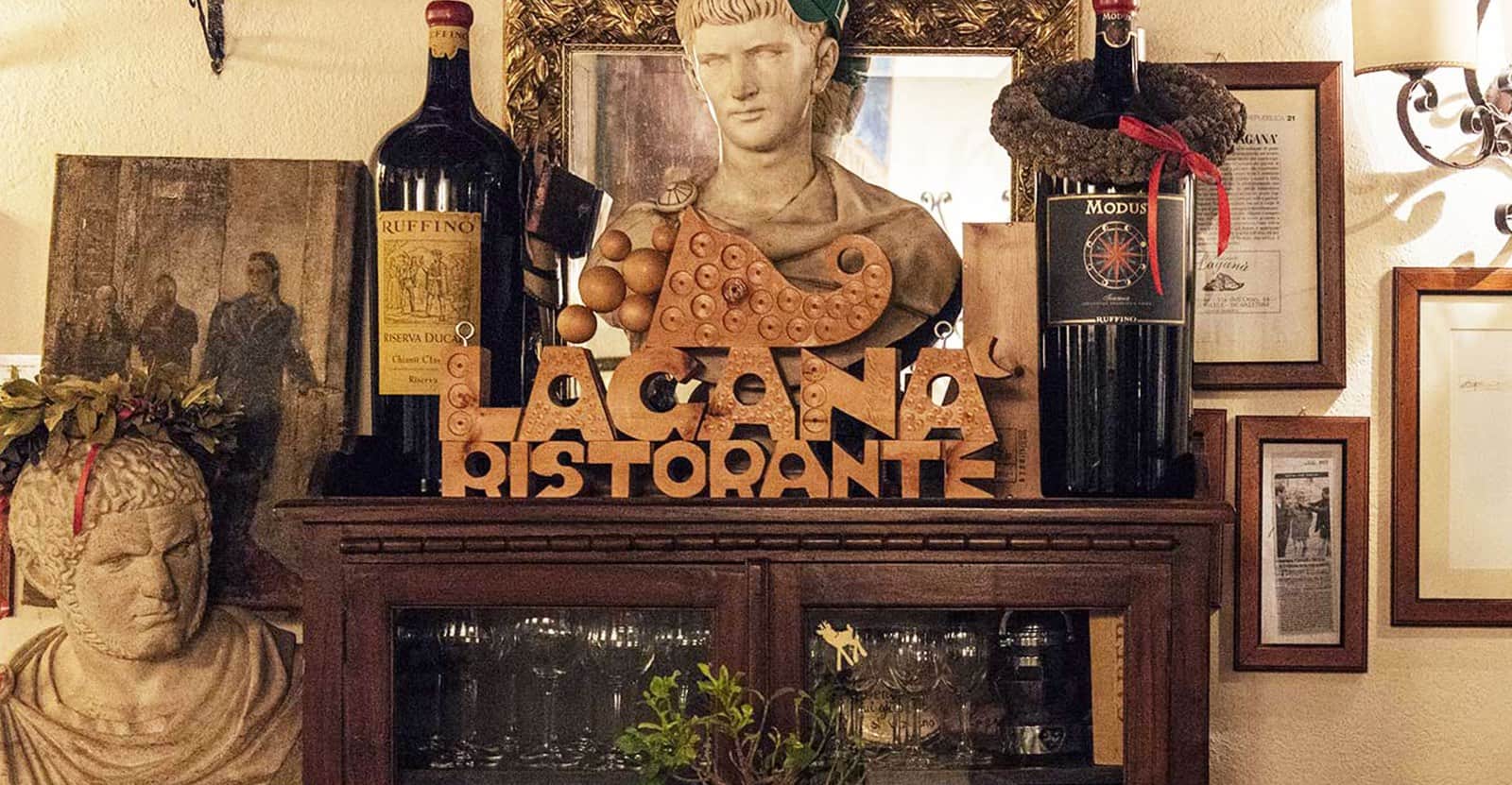 Insegna in legno con scritta Laganà Ristorante su un mobile, tra bottiglie di vino e un busto romano.