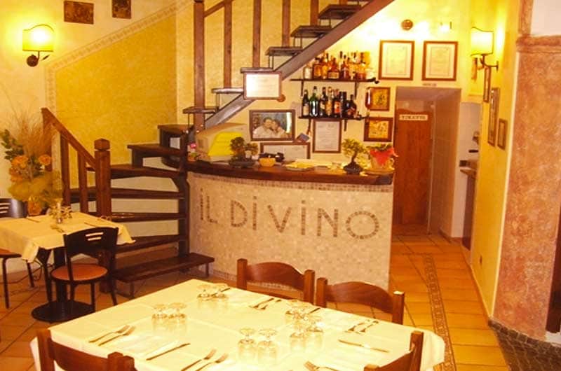 Sala con scala in legno, bancone rivestito a mosaico con la scritta IL DIVINO e tavoli apparecchiati in primo piano.
