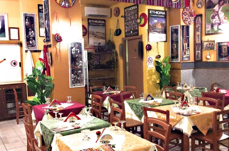 Sala da ristorante con pareti gialle coperte di poster e maschere, tavoli con tovaglie verdi e bordeaux apparecchiati.