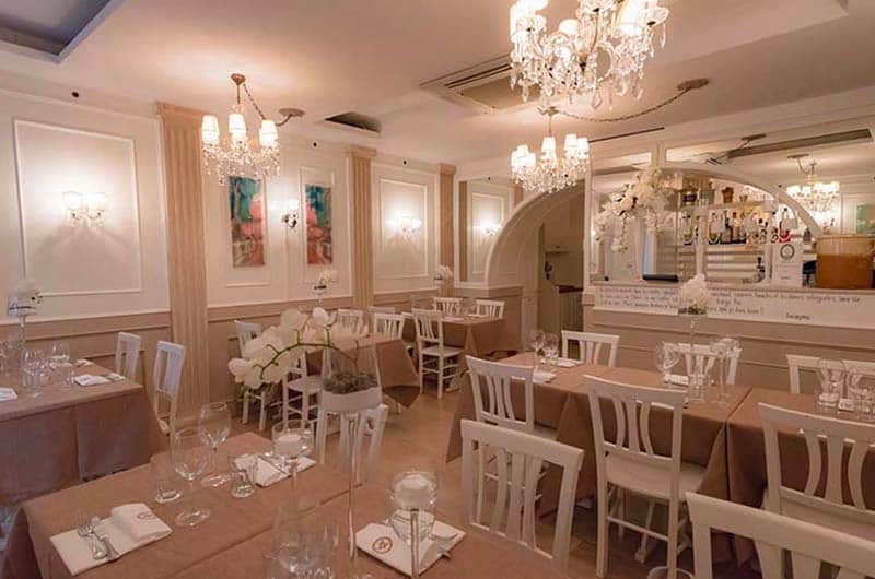Sala da ristorante con tavoli apparecchiati in tonalità rosa cipria, sedie bianche e lampadari di cristallo.
