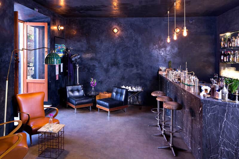Sala di un bar con bancone in marmo scuro, sgabelli in legno e poltrone in pelle sotto pareti scure.