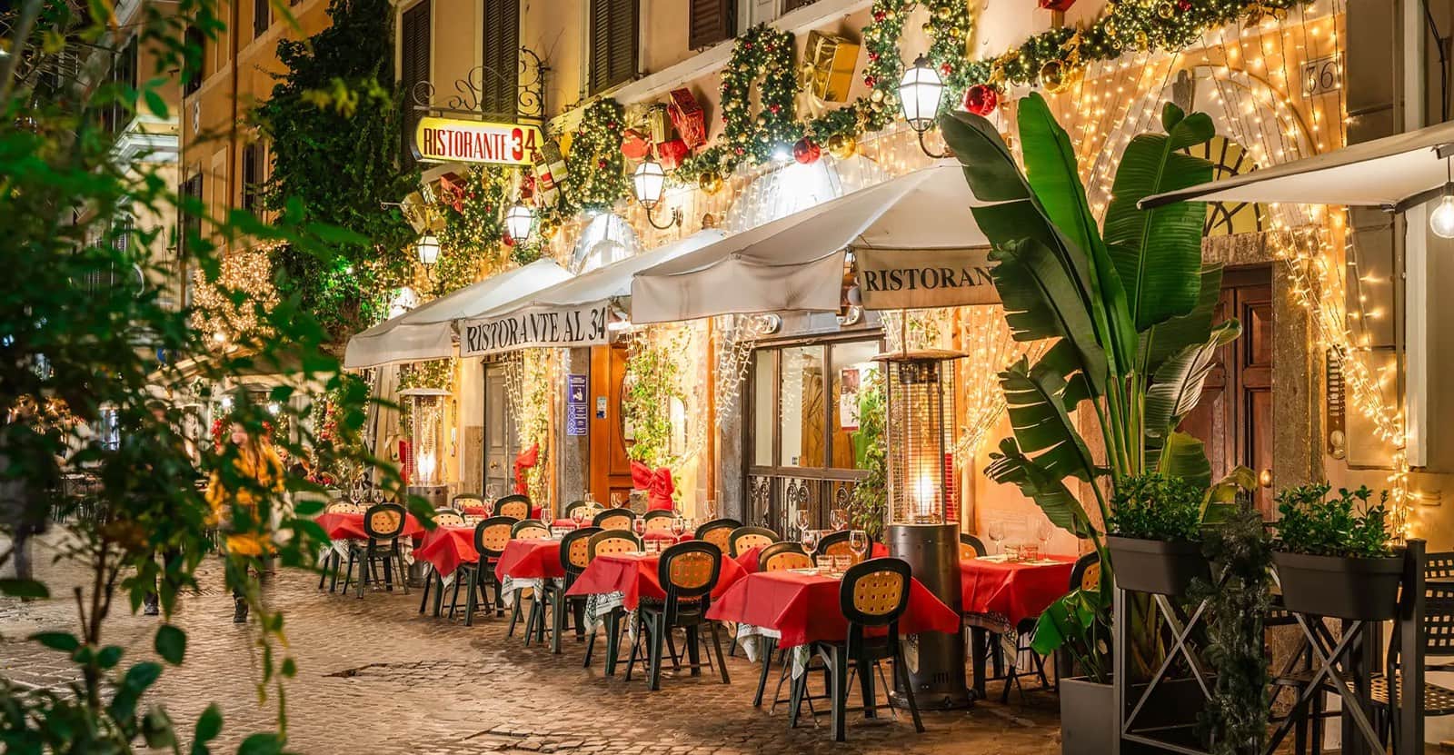Tavoli con tovaglie rosse su un vicolo acciottolato, sotto tende bianche e decorazioni natalizie luminose.